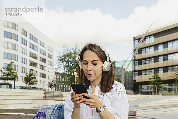 Lächelnde Frau hört Musik über kabellose Kopfhörer und benutzt Smartphone auf Bank