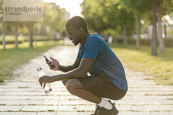 Glücklicher Mann kauert auf Fußweg und benutzt Smartphone im Park