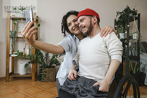 Lächelnde Frau mit Freund im Rollstuhl  die zu Hause ein Selfie mit dem Handy macht