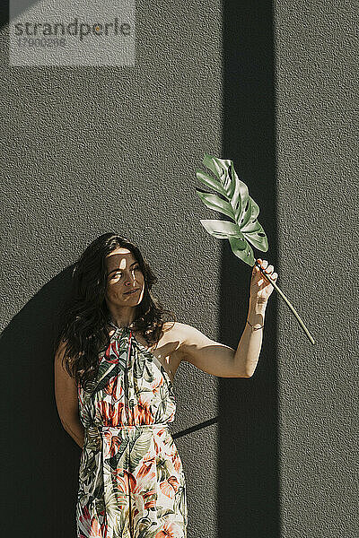 Woman shielding face with monstera leaf in front of wall on sunny day