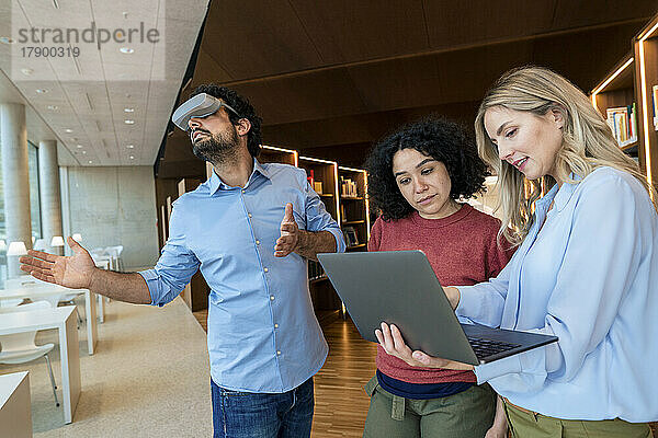 Zwei Frauen arbeiten am Laptop von einer Kollegin mit VR-Brille in der Bibliothek