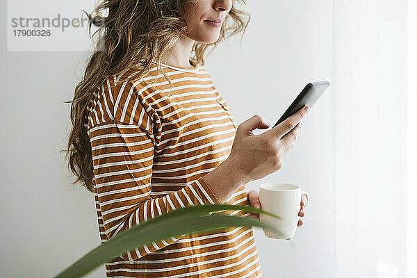 Junge Frau hält Kaffeetasse in der Hand und benutzt Mobiltelefon