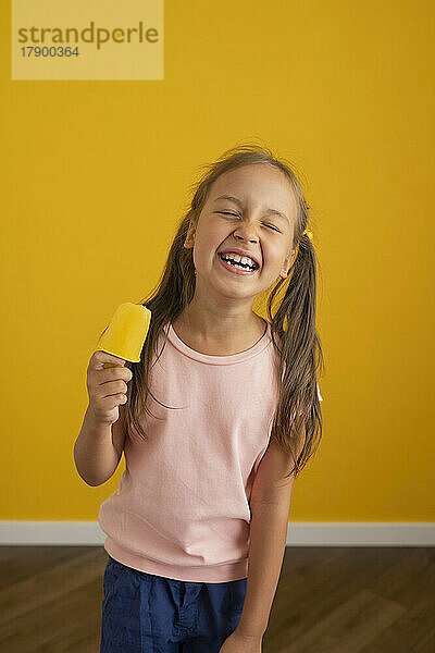 Happy girl standing with ice cream in front of yellow wall