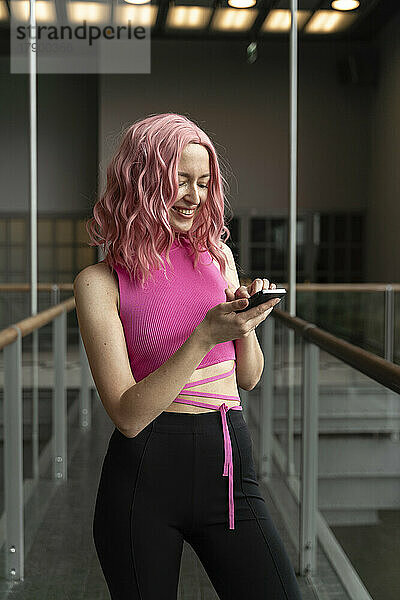 Lächelnde Frau mit rosa Haaren surft im Flur über ihr Smartphone im Internet