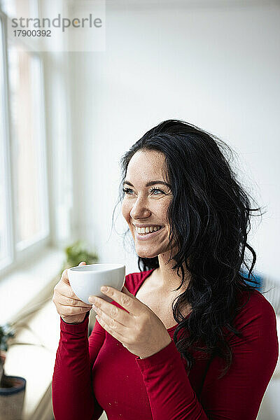 Glückliche Frau mit Kaffeetasse und Blick durch das Fenster