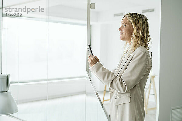 Geschäftsfrau hält Mobiltelefon im minimalistischen Bürokorridor