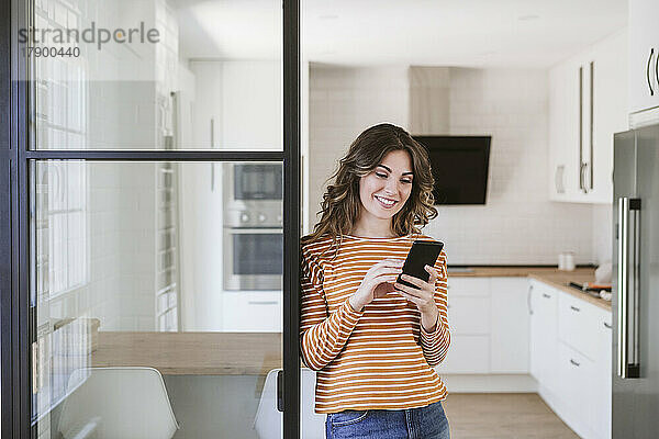 Lächelnde junge Frau benutzt ihr Mobiltelefon an einer Glaswand in der Küche