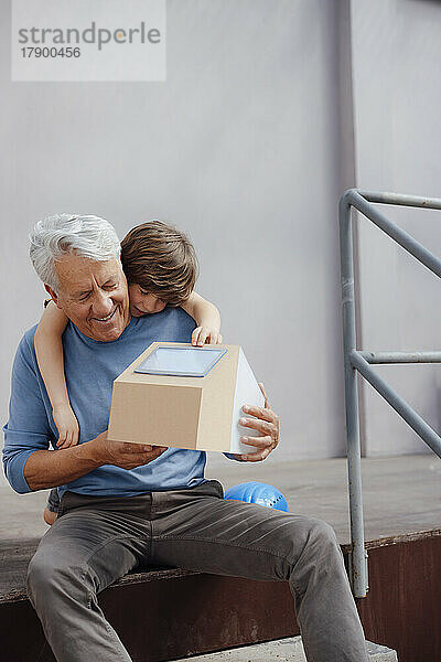Lächelnder älterer Mann hält das Solarhausmodell seines Enkels vor der Wand