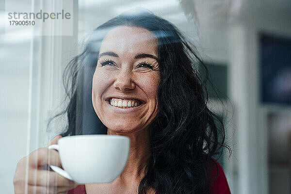 Fröhliche Frau mit Kaffeetasse durch Glas gesehen
