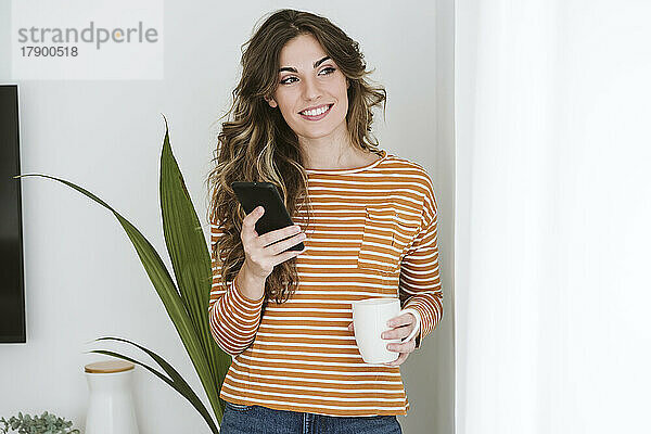 Lächelnde junge Frau mit Kaffeetasse und Mobiltelefon