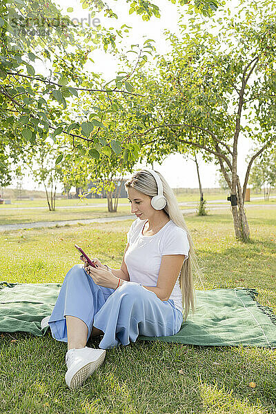 Junge Frau mit kabellosen Kopfhörern und Smartphone sitzt im Park