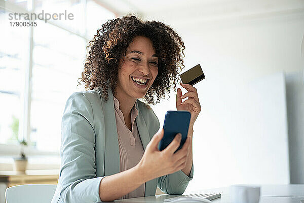 Glückliche Geschäftsfrau  die am Arbeitsplatz online per Smartphone bezahlt