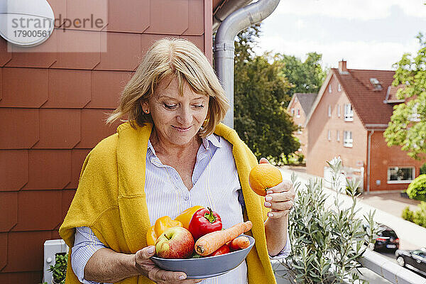 Lächelnde Frau mit einer Schüssel mit frischem Obst und Gemüse auf dem Balkon