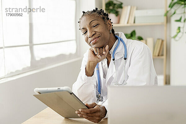 Lächelnder Arzt mit der Hand am Kinn  der im Heimbüro am Tisch sitzt
