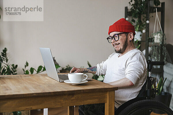 Lächelnder Mann im Rollstuhl  der zu Hause Laptop benutzt