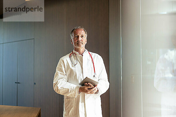 Selbstbewusster Oberarzt hält Tablet-PC in der Nähe der Glaswand im Krankenhaus