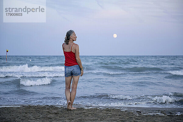 Frau steht am Strand mit Mond über dem Horizont