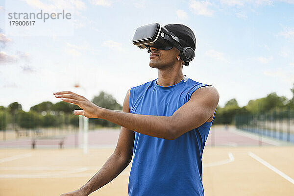 Junger Mann mit VR-Brille gestikuliert stehend auf dem Basketballplatz