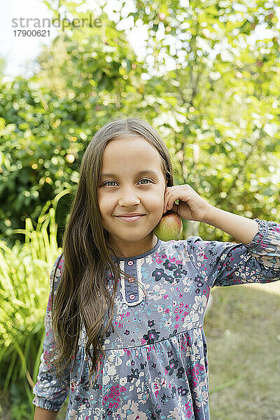 Lächelndes Mädchen hält Apfel am Ohr im Garten