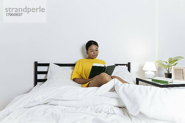 Frau im gelben T-Shirt sitzt auf dem Bett und liest ein Buch