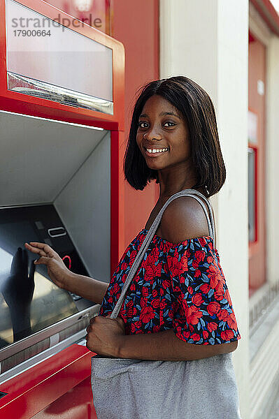 Lächelnde Frau mit Umhängetasche steht am Geldautomaten