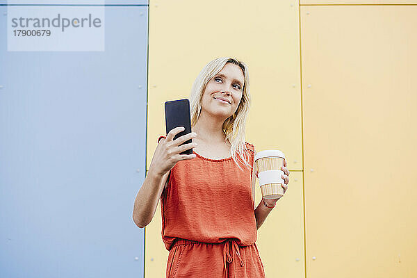 Lächelnde junge Frau mit Mobiltelefon und Einwegkaffeetasse vor bunter Wand