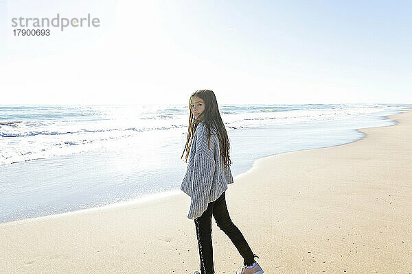 Mädchen trägt Pullover und steht an einem sonnigen Tag am Strand