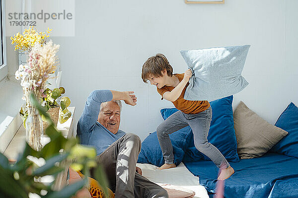 Fröhlicher Junge spielt Kissenschlacht mit Großvater auf dem heimischen Sofa