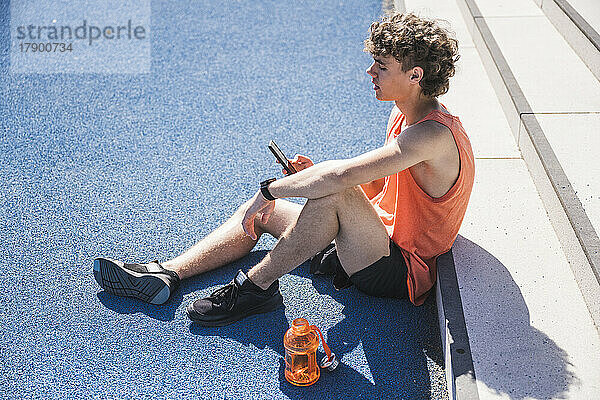 Junger Sportler sitzt an einem sonnigen Tag mit seinem Smartphone neben den Stufen