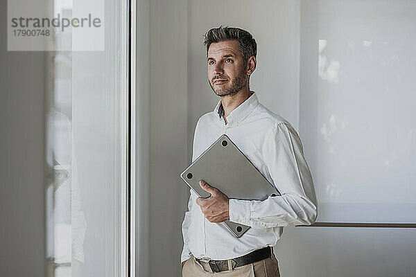 Nachdenklicher Geschäftsmann mit Laptop und Blick durchs Fenster steht mit der Hand in der Tasche im Büro