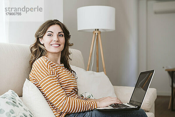 Lächelnde junge Frau  die zu Hause auf dem Sofa einen Laptop benutzt