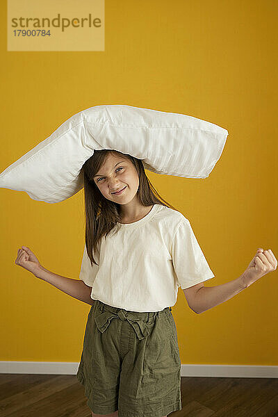 Lächelndes Mädchen  das ein weißes Kissen auf dem Kopf trägt und Fäuste zeigt