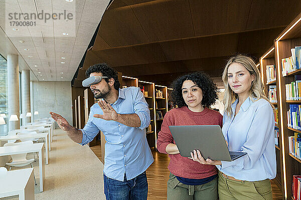 Zwei Frauen mit Laptop von einer Kollegin  die durch eine VR-Brille in der Bibliothek zuschaut