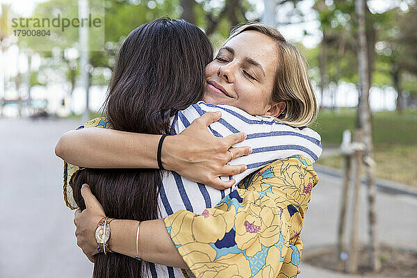 Lächelnde Frau mit geschlossenen Augen umarmt Freundin im Park