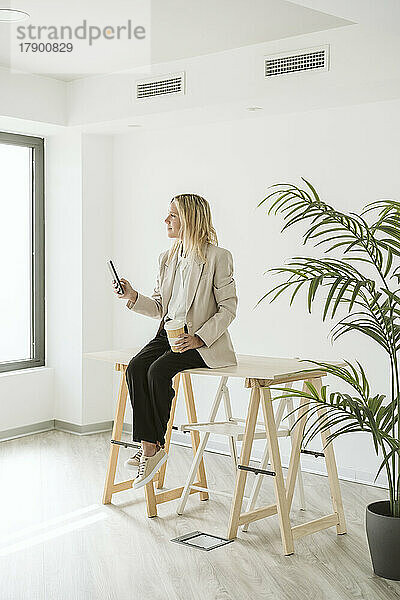 Geschäftsfrau sitzt am Schreibtisch im modernen Büro und hält ihr Mobiltelefon in der Hand