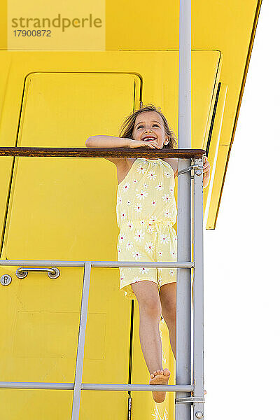 Smiling girl standing on railing in front of yellow lifeguard hut