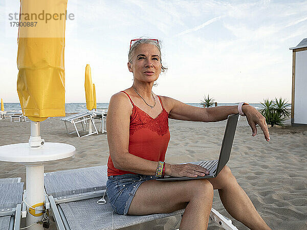 Nachdenkliche ältere Geschäftsfrau sitzt mit Laptop am Strand