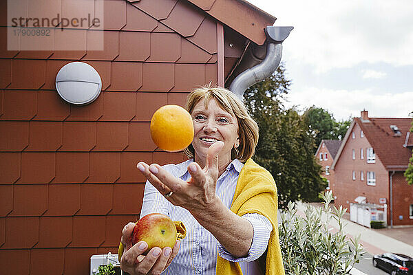 Glückliche Seniorin jongliert mit Orangen- und Apfelfrüchten auf dem Balkon