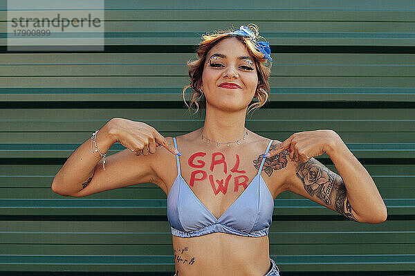 Selbstbewusste Frau zeigt Girl-Power-Text auf der Brust vor grüner Metallwand