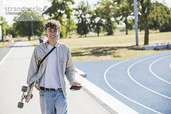 Glücklicher junger Mann mit Skateboard und Mobiltelefon auf dem Fußweg