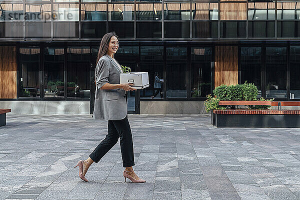 Lächelnde Geschäftsfrau  die mit einem Karton vor dem Bürogebäude läuft