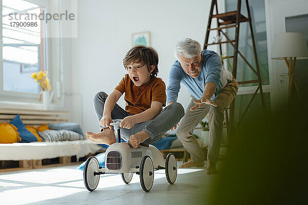 Cheerful grandfather playing with grandson sitting on toy car in living room