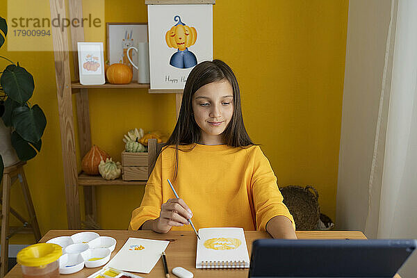 Lächelndes Mädchen malt auf einem Skizzenblock und schaut sich zu Hause ein Online-Tutorial über einen Tablet-PC an