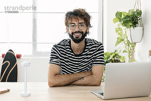 Lächelnder Mann sitzt neben Windkraftanlage und Laptop im Heimbüro