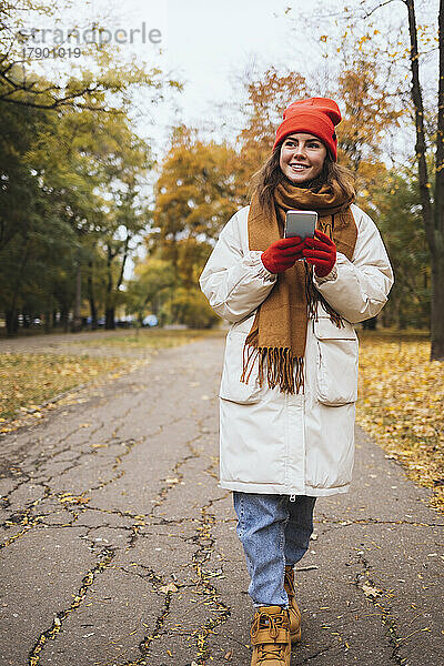 Lächelnde junge Frau mit Mobiltelefon geht auf Fußweg im Park