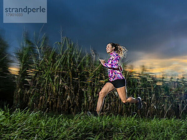Smiling young sportswoman running on grass at dusk
