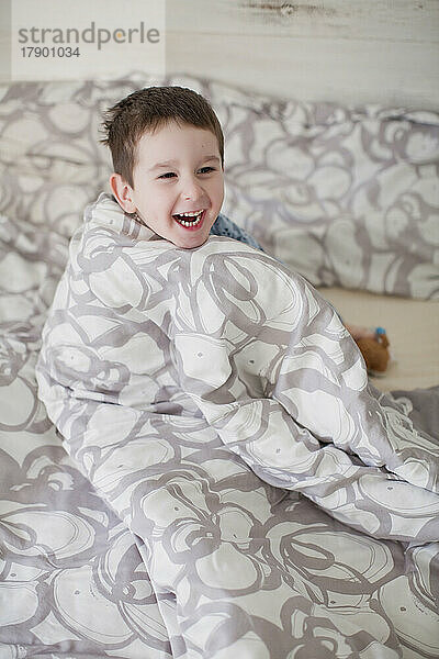 Fröhlicher Junge  in Decke gehüllt auf dem Bett