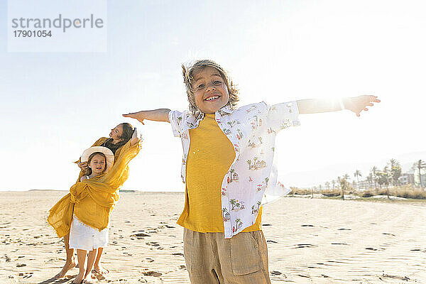 Fröhlicher Junge mit ausgestreckten Armen steht an einem sonnigen Tag am Strand