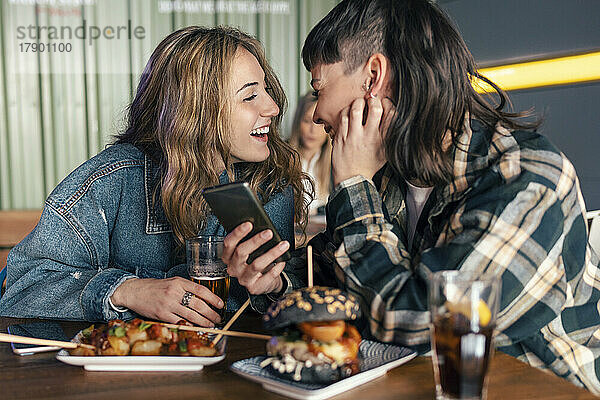 Liebevolles lesbisches Paar mit Smartphone sitzt am Tisch im Restaurant