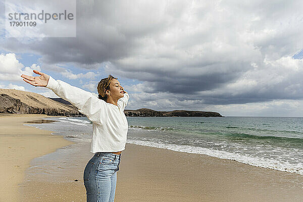 Junge Frau mit ausgestreckten Armen und geschlossenen Augen genießt den sonnigen Tag am Strand